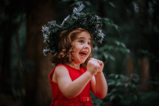 A joyful child wearing a festive wreath and bright red dress, expressing excitement outdoors.