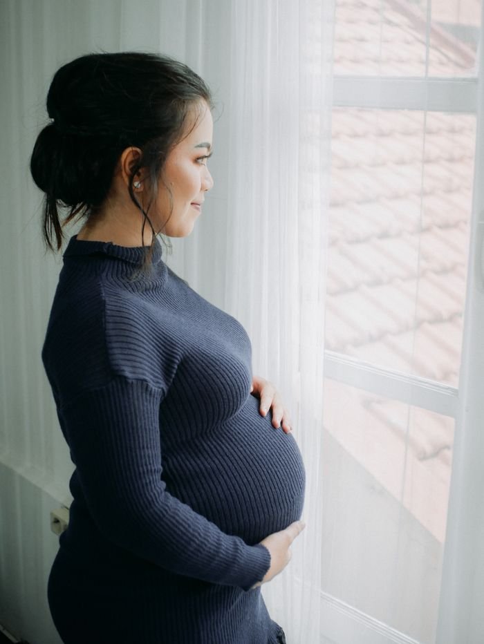 finding-rest-in-god Pregnant woman in a blue sweater looking out a window, embracing motherhood in quiet reflection, trusting God.