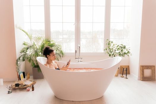 a woman in a bathtub enjoying a book surrounded by plants and nice outdoor windows overlooking the yard. 